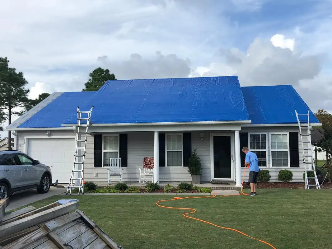 A person stands on the lawn in front of a single-story house with a blue tarp covering the roof, presumably for a roof replacement in Sarasota County. Two ladders are placed against the house, and an orange hose is laid out on the ground. An SUV is parked in the driveway, and the sky is partly cloudy.