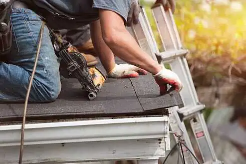 A worker wearing gloves is using a power tool to install shingles on a roof, likely during a roof replacement in Sarasota County. Another worker's hands are visible, steadying a ladder nearby. The scene is set against a blurred background, possibly showing greenery and sunlight.