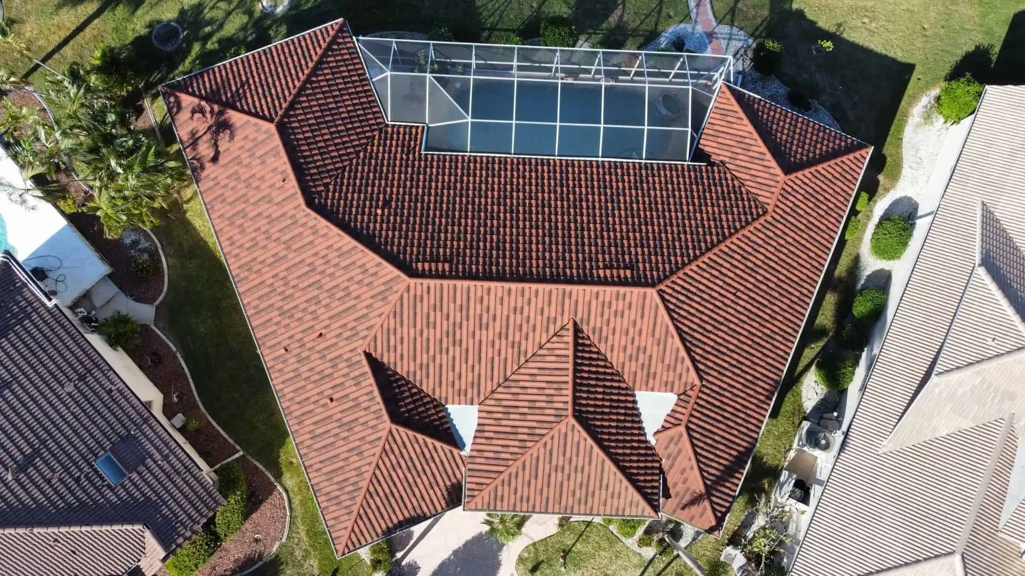 Aerial view of a house with a complex red-tiled roof and an enclosed outdoor area featuring a swimming pool. The property is surrounded by green lawns and landscaped gardens, with neighboring homes on either side—a perfect candidate for metal roof replacement by experts in Sarasota County.