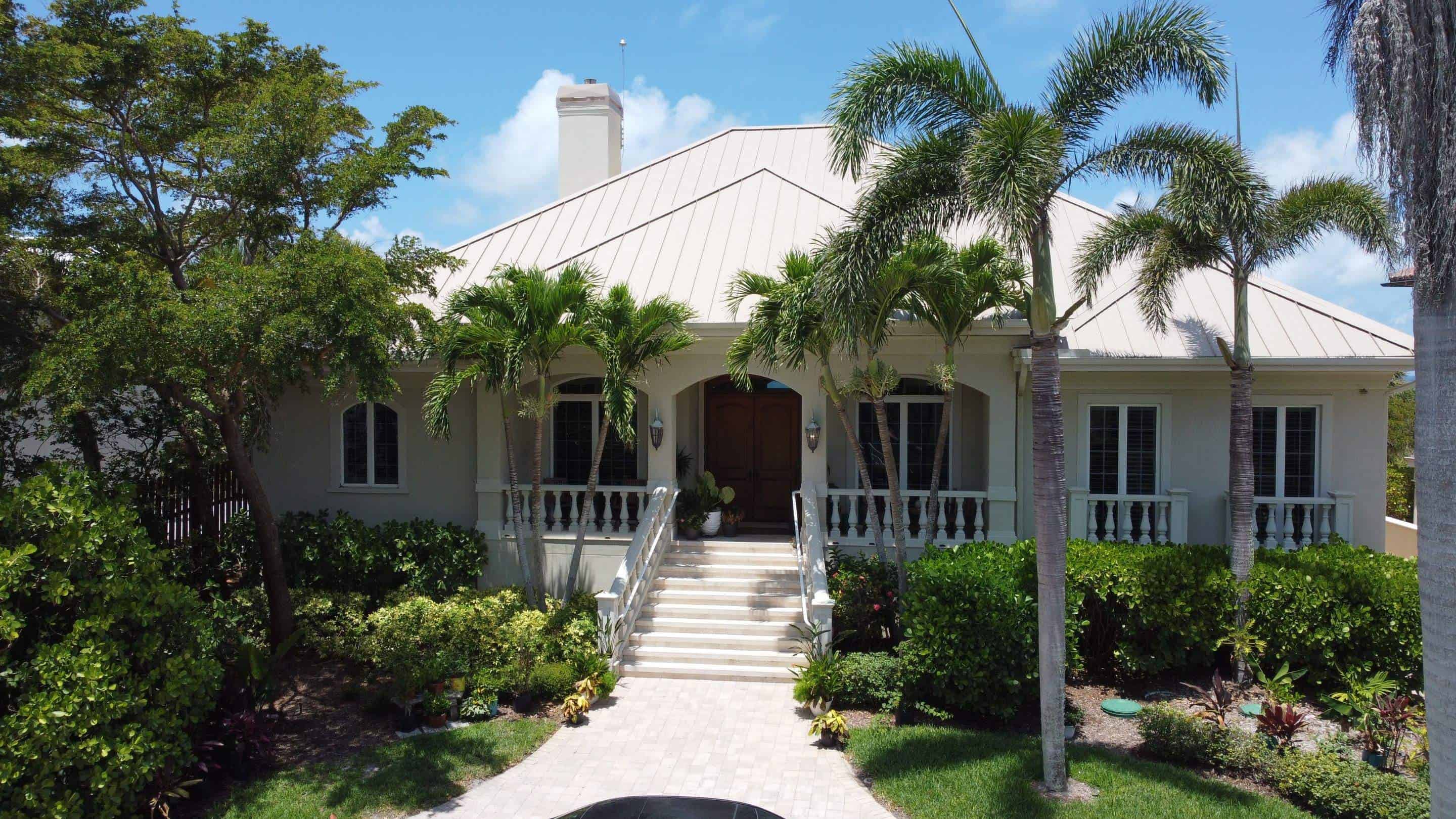 A tropical-style house with a metal roof is surrounded by lush greenery. Palm trees frame the entrance, which features white steps leading up to a covered porch with arches and wooden double doors, all crafted by a renowned roofing contractor from Sarasota County. The sky is clear and blue in the background.