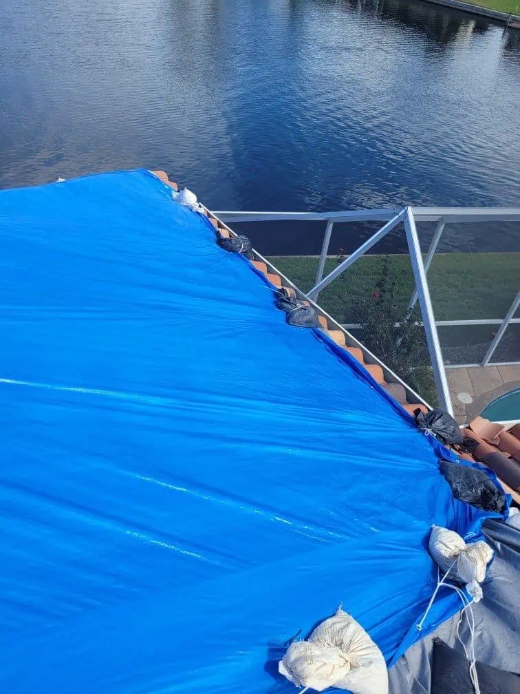 A rooftop covered with a blue tarp, secured with sandbags, overlooks a body of water. The roof appears to be undergoing Roof Replacement Sarasota County. In the background, there is a fenced-in area and a pool visible to the right.
