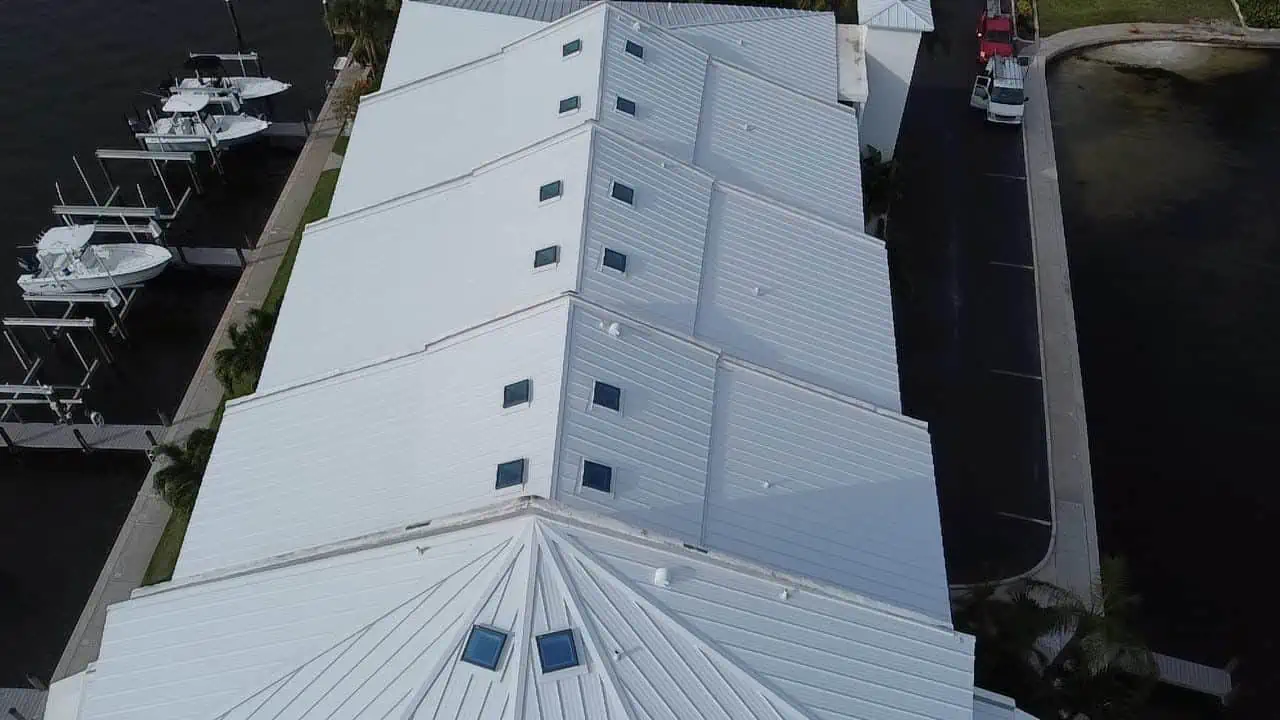 Aerial view of a large white metal-roofed building with multiple triangular roof sections and small windows. The building, which may have been serviced by a roofer in Sarasota County, is adjacent to a marina with docked boats on one side and a parking area with parked vehicles on the other. The image is taken during the day.