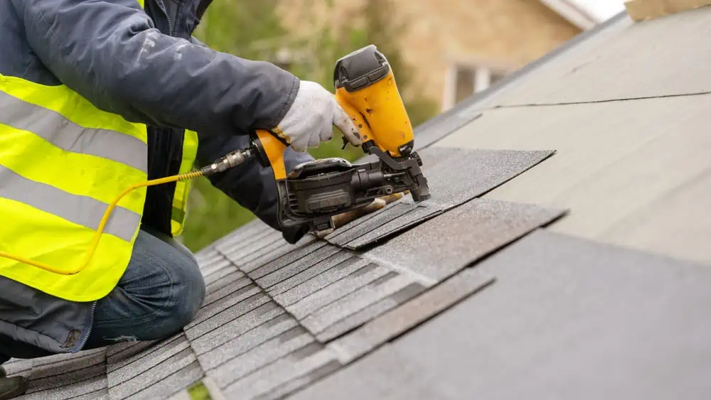 A worker in a high-visibility vest, a Roofer in Sarasota County, uses a pneumatic nail gun to install asphalt shingles on a sloped roof. The worker's hands and tools are shown in close-up, highlighting the action of fastening the shingles. Trees and a house are blurred in the background.