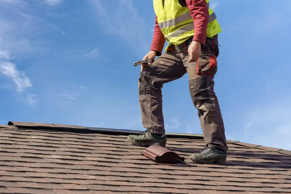 A construction worker, wearing a yellow safety vest and holding a tool, stands on a shingled roof under a clear blue sky. The worker, likely from Roofing Contractor Sarasota County, is preparing to install or repair a section of the roof.
