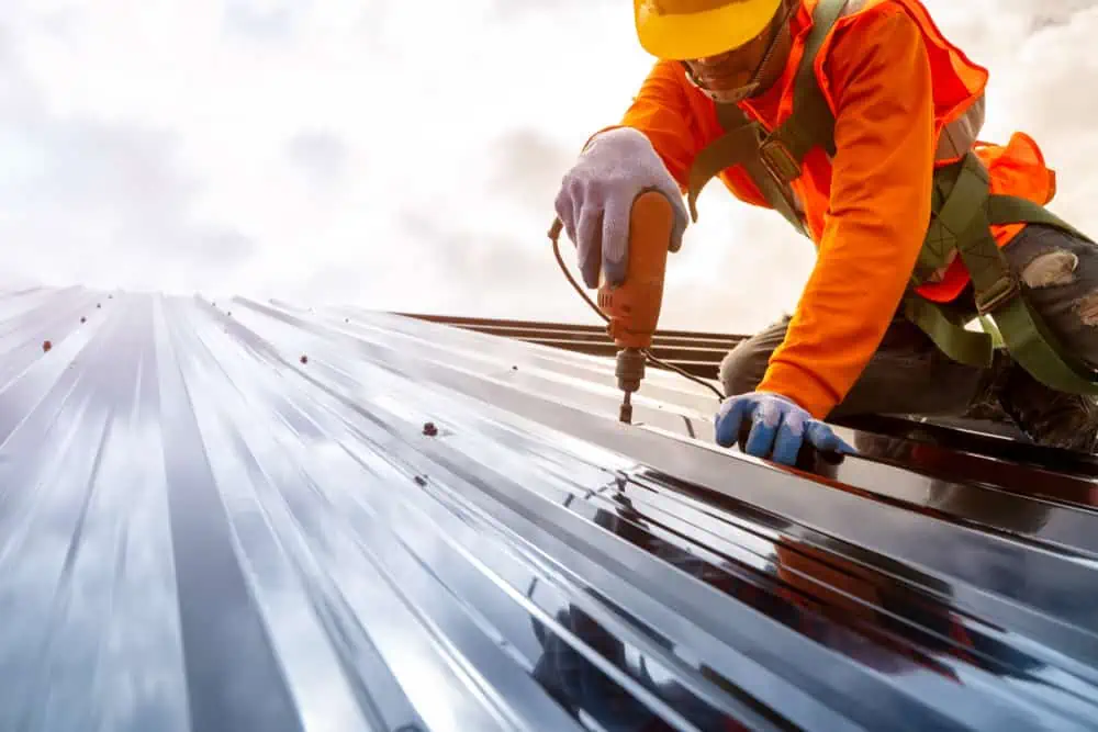 A worker wearing an orange safety jacket, yellow hard hat, and gloves is using a power tool to secure a metal sheet on a roof. The worker is kneeling and focused on the task, perhaps part of Roof Replacement Sarasota County, with structural beams and a cloudy sky in the background.