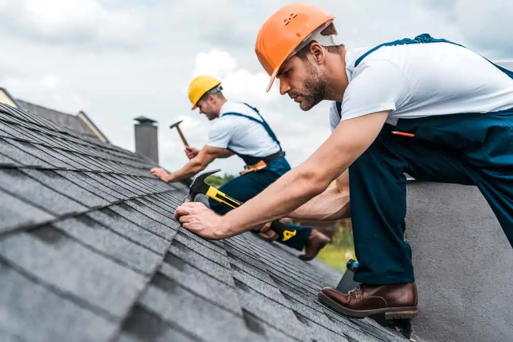 Two construction workers wearing hard hats and safety gear are working on a house roof. One in the foreground hammers shingles down while another in the background attends to additional tasks. Both are on a gray tiled roof under a cloudy sky, showcasing their expertise as a roofing contractor in Sarasota County.