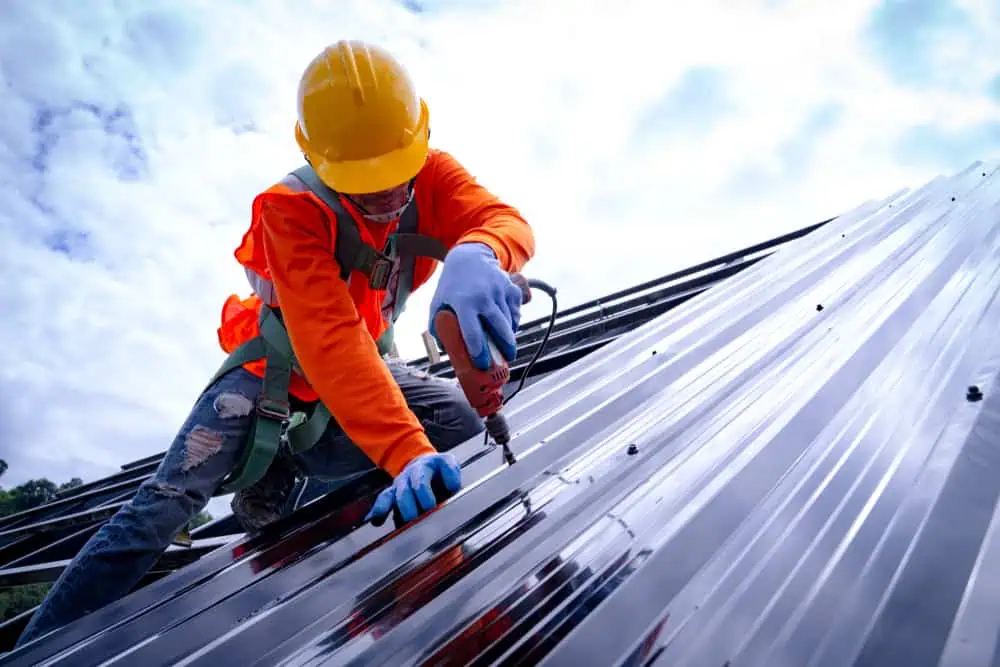 A worker wearing a yellow hard hat and orange safety vest is using a power tool to install or repair a metal roof in Sarasota County. Secured with a safety harness, the worker focuses on the roof replacement as the sky remains overcast in the background.