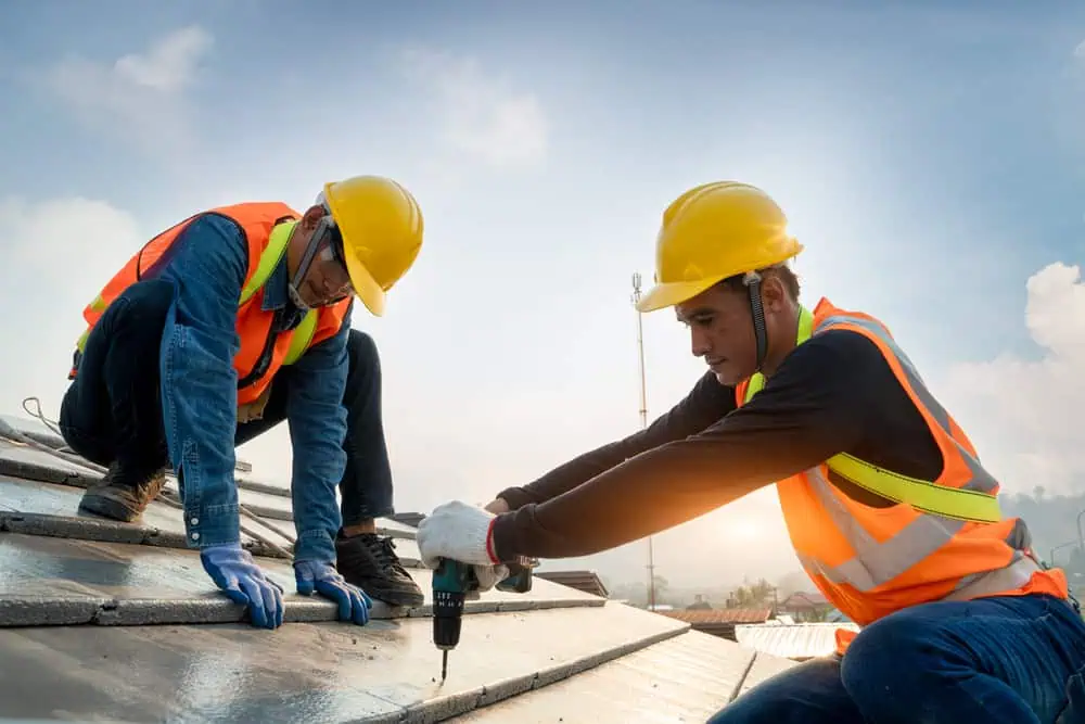 Two construction workers wearing safety vests and helmets are working on a metal roof in Sarasota County. One worker is using a drill to secure roofing material while the other is holding it in place. The sky in the background is clear with a hint of clouds.