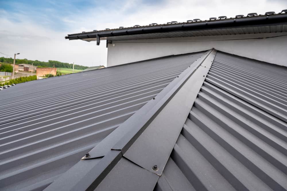 A close-up of a modern, dark gray metal roof with ridges and seams. The roof has secure fastening points along the seams, ideal for a thorough Roof Inspection Sarasota County. The sky is partly cloudy in the background, showing part of a building's exterior wall and nearby surroundings.