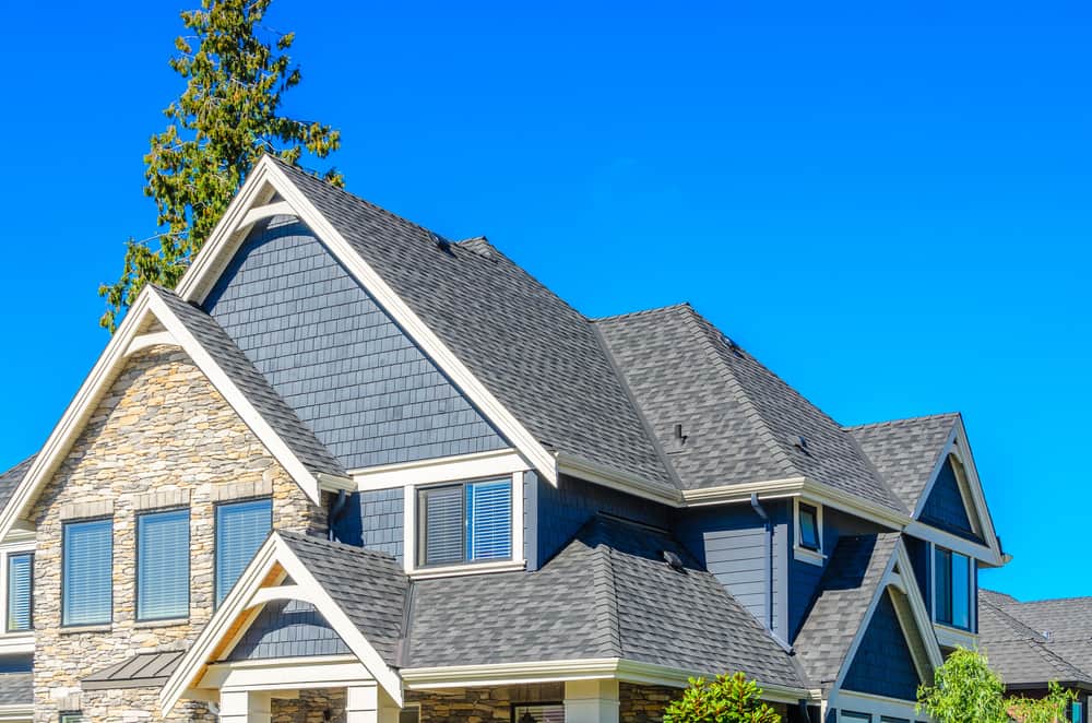 A two-story house with a dark grey roof, blue-grey siding, stone accents, and white trim. The house boasts multiple gables and windows with blue shutters. There is a tall evergreen tree and a clear blue sky in the background, all maintained by a top-notch roofing contractor from Sarasota County.