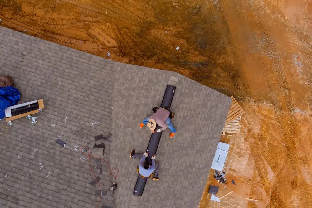 Aerial view of two workers installing a vent on a shingled roof. The surrounding area shows construction materials and dirt, indicating an ongoing building project by a roofing contractor in Sarasota County. One worker is handling the vent while the other is assisting and holding the structure steady.