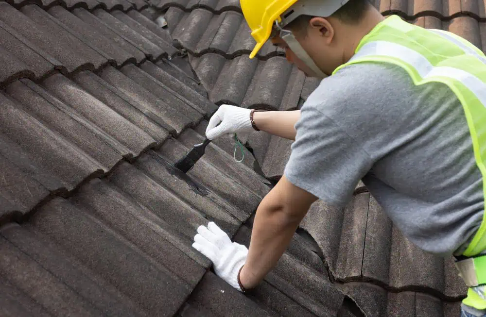 A person wearing a yellow hard hat, safety vest, gray shirt, and white gloves is expertly repairing a roof in Sarasota County. They meticulously apply a substance to the overlapping dark gray tiles using a brush. The roofing contractor is focused on their task to ensure quality results.