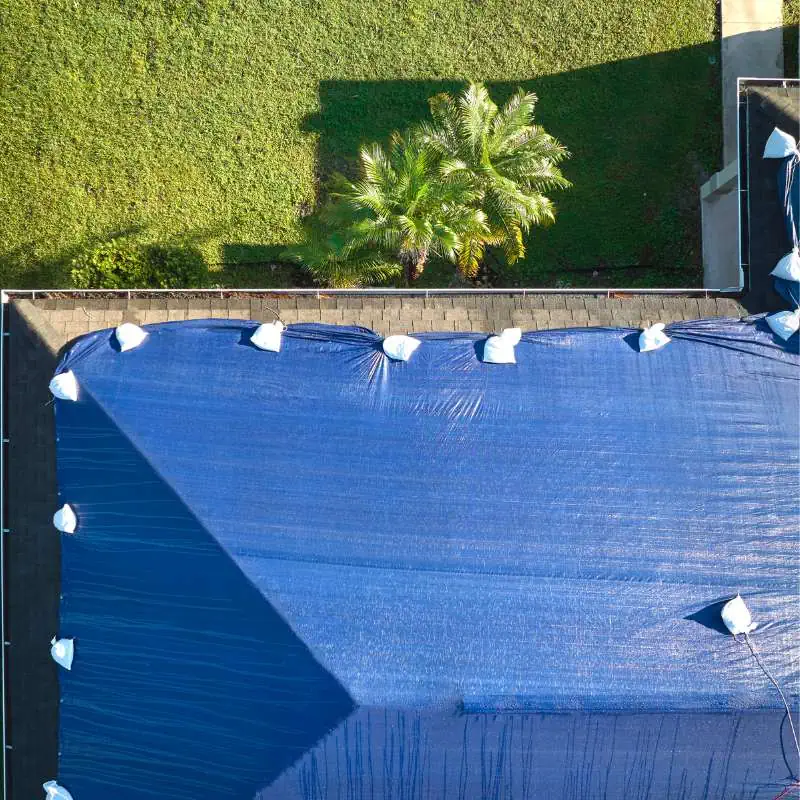 Aerial view of a blue tarp spread over a roof with white sandbags anchoring its edges. To the side, there is a grass lawn with a few palm trees casting shadows. The tarp signals an ongoing roof replacement in Sarasota County, and the top edge aligns partially with the building beside a paved area.