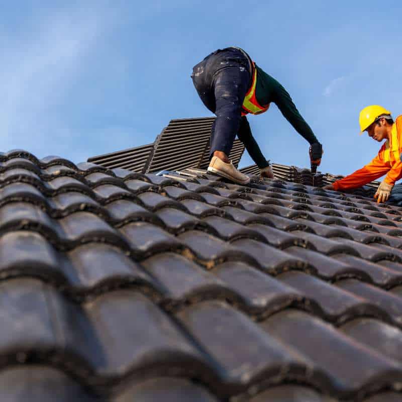 Two workers wearing safety gear and hard hats are seen installing roof tiles on a sloped roof in Sarasota County. One worker is crouching while the other is standing, both focused on the task. The clear blue sky forms a serene backdrop, ideal conditions for a thorough roof inspection in Sarasota County.