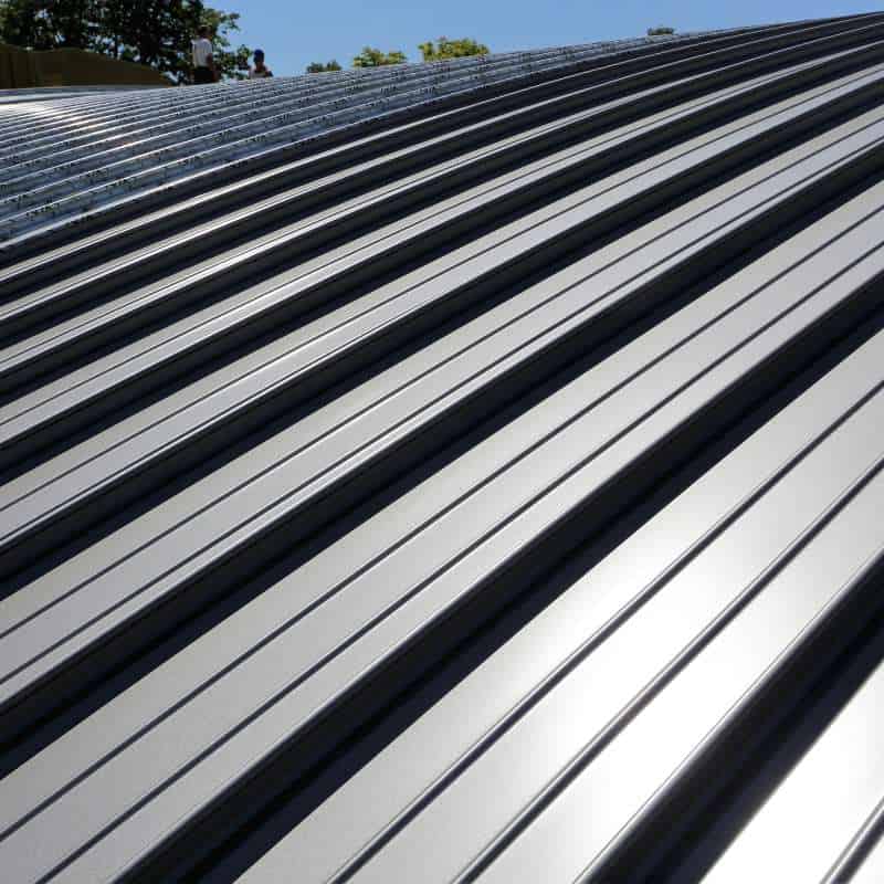 Close-up view of a modern metal roof with parallel, evenly spaced ridges running down its surface. The metallic panels reflect sunlight, showcasing a sleek and polished finish. A clear blue sky and some tree branches are visible in the background—ideal work by a reputable Roofer Sarasota County.