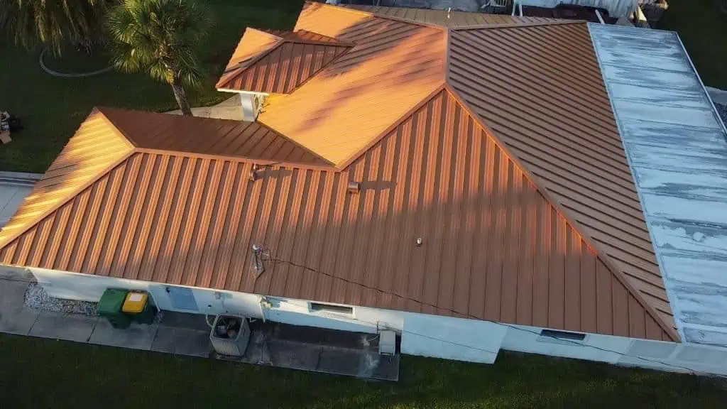 Aerial view of a house with multiple pitched metal roofs in a reddish-brown color. A small palm tree stands near the house, with various objects including trash bins and storage containers visible on the property. If you're considering roofing companies Sarasota County, note the adjacent roof is partially visible.