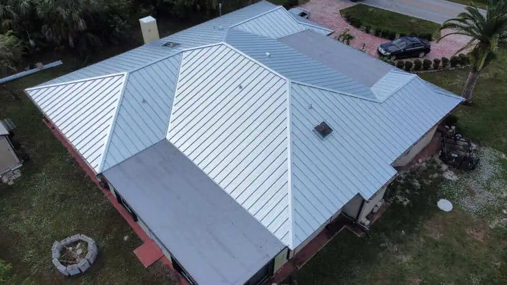 Overhead view of a large house with a modern, silver-colored metal roof installed by a top roofer in Sarasota County. The roof features multiple angular sections. The surrounding yard has a fire pit area, paved driveway, and some trees and shrubs.