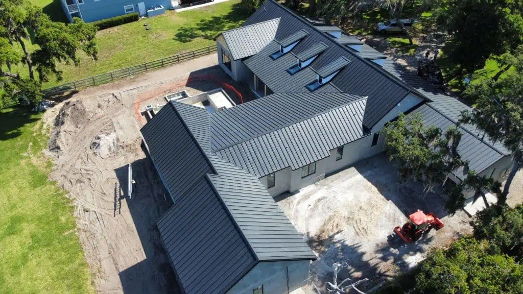 Aerial view of a large, partially constructed building with a metal roof. The surrounding area features dirt mounds and construction equipment. Trees and grassy fields can be seen around the property, indicating a rural setting where roofing contractors in Sarasota County are hard at work.
