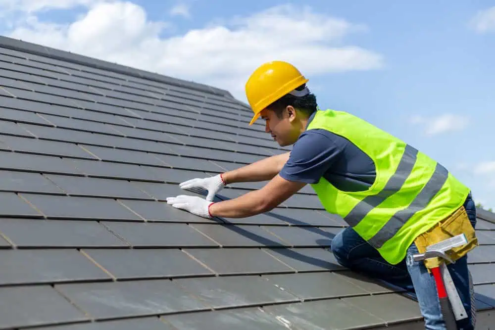 A construction worker wearing a yellow hard hat, neon yellow safety vest, and gloves is repairing or installing roof shingles on a sloped roof. The sky is clear with a few clouds in the background, ideal for a Roof Inspection Sarasota County to ensure everything is in top shape.