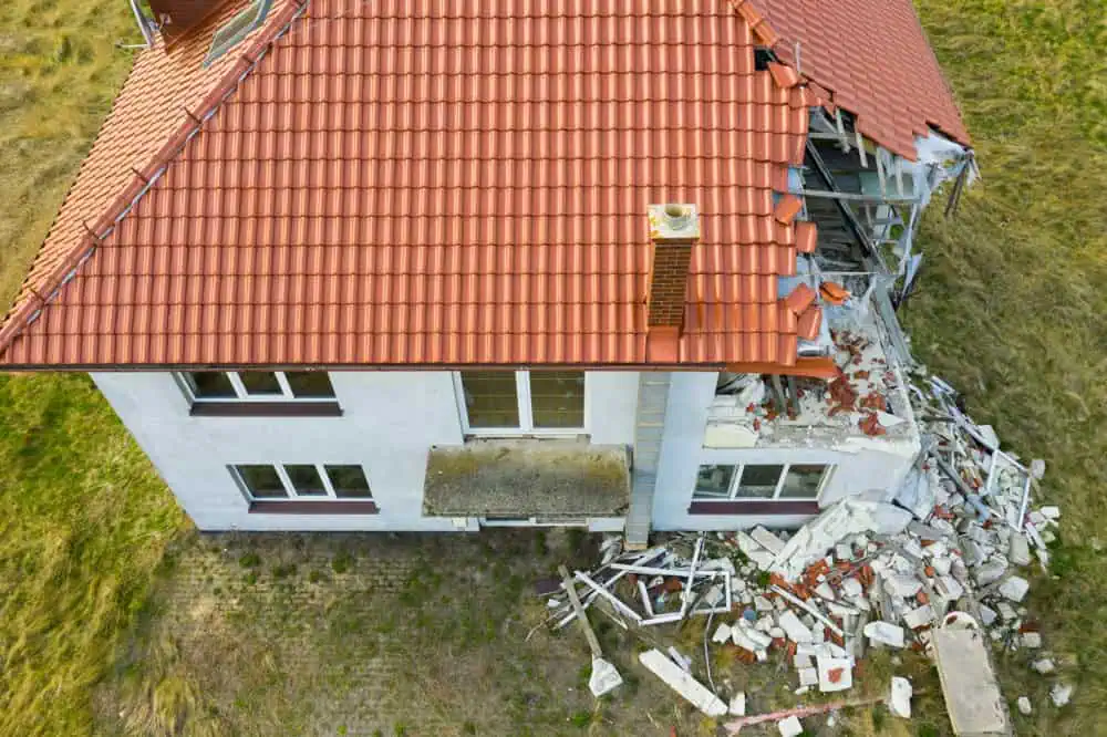 An aerial view of a two-story house with a red tiled roof in Sarasota County shows significant structural damage. One corner of the roof is severely collapsed, exposing interior spaces and scattered debris on the ground. Surrounding the house are grassy areas, indicating an urgent need for roof repair in Sarasota County.