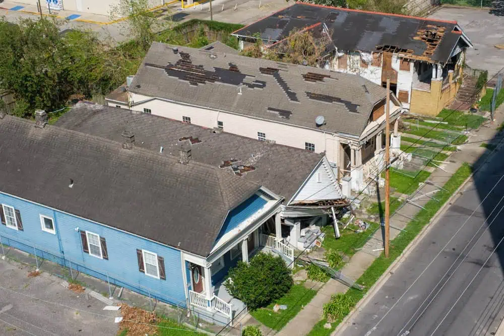 Aerial view of three distressed houses showing significant roof damage, one painted blue and the others beige, located along a street in Sarasota County. The surrounding area is littered with debris and overgrown vegetation, indicating a dire need for roof repairs or replacements.