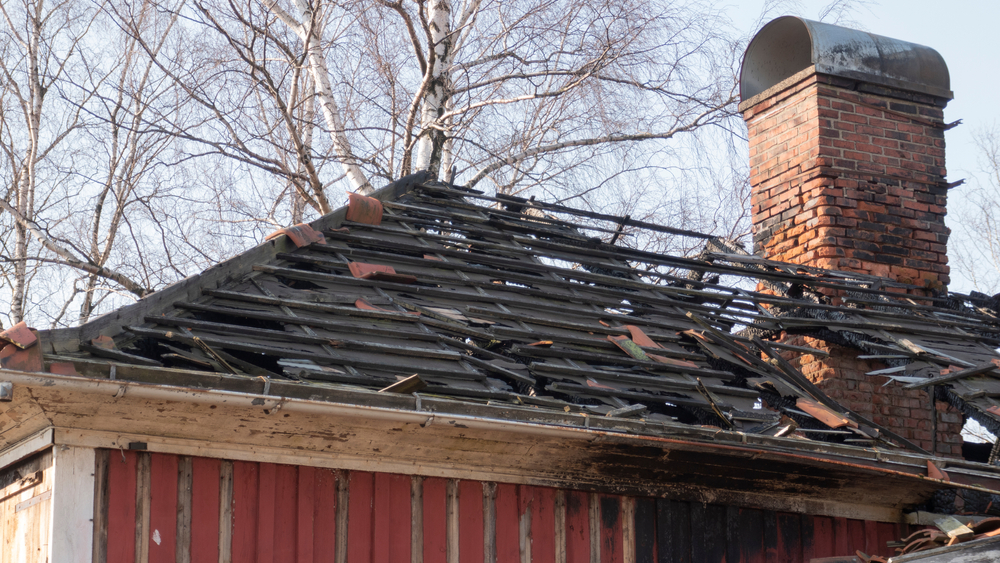 A red wooden house with a severely damaged roof missing shingles, exposing the wooden structure. The chimney remains intact. Leafless trees are visible in the background, under a clear blue sky.