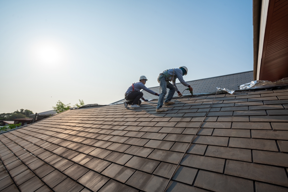 Two workers wearing safety gear are installing shingles on a sloped roof under a clear blue sky. Sunlight is visible in the background, and the surrounding landscape can be seen in the distance.