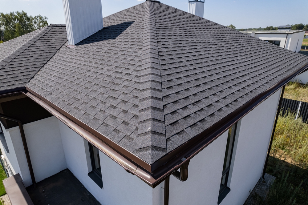 A house with a grey, shingled hip roof is shown in close-up. The building has white walls and a small flat section beneath the roof. Two chimney structures are visible. Greenery surrounds the house in the background.