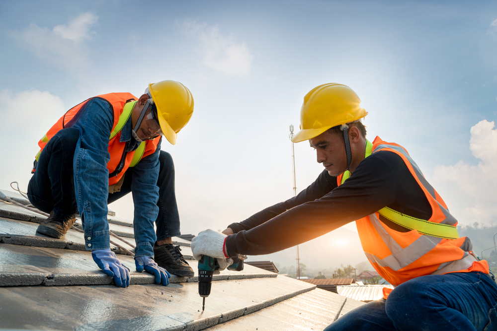 Two construction workers in safety vests and helmets are installing solar panels on a roof. One is drilling while the other secures the panels, under a clear sky.