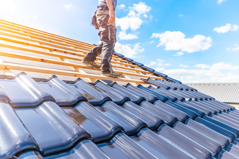 A construction worker installs dark blue ceramic tiles on a rooftop under a partly cloudy sky. The sun shines brightly, casting reflections on the tiles. The worker is wearing work pants and boots.