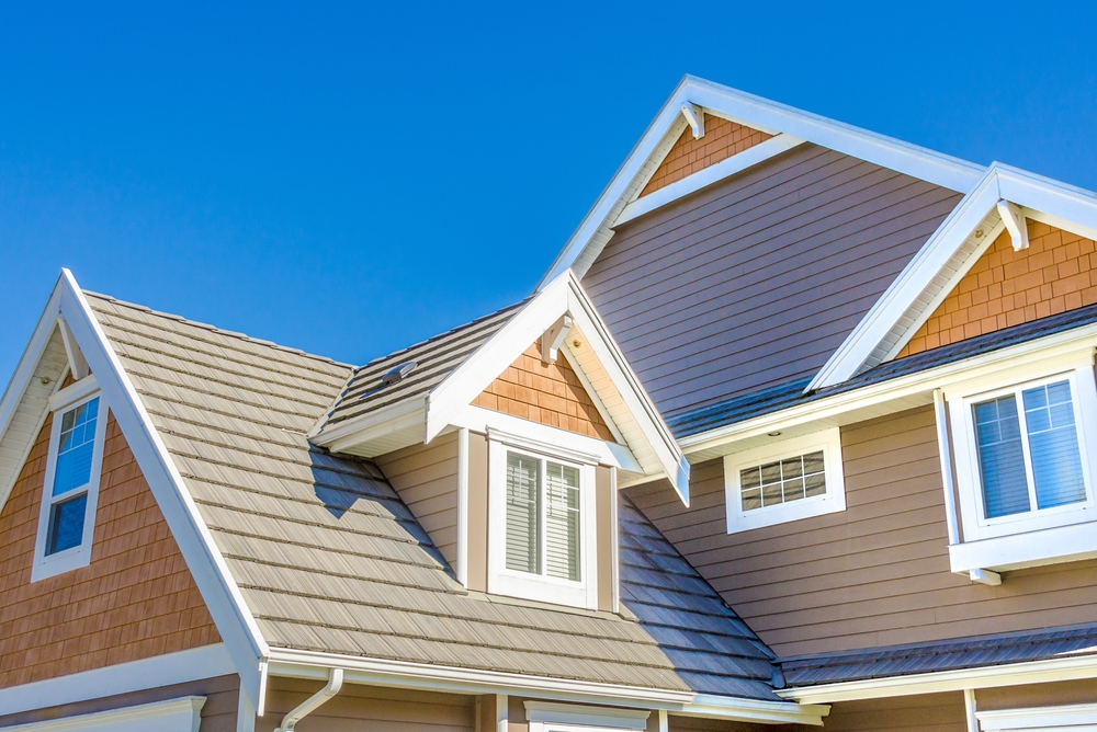 The image shows the roofline of a modern suburban house with brown and beige siding and white trim. The sky is clear and blue, providing a sharp contrast to the home's exterior. The architecture features multiple gables and windows.