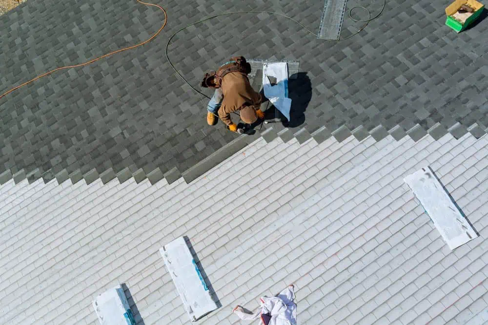 A worker in brown overalls and a helmet installs gray shingles on a roof, with tools and materials around. The roof is partially covered with white underlayment. An orange extension cord and a green box are visible on the sides.