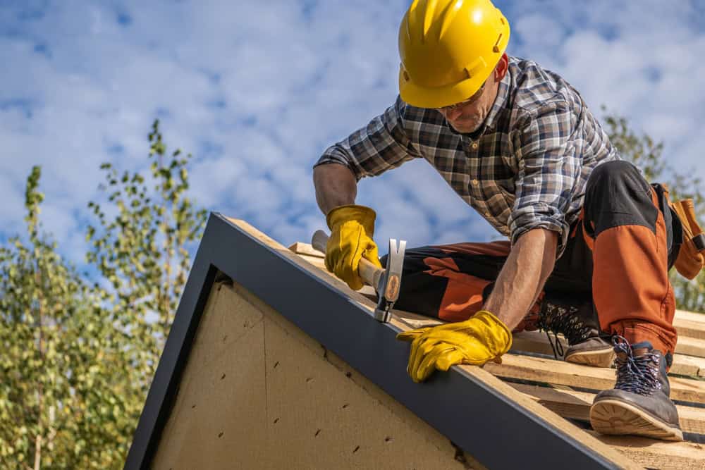 A construction worker in a yellow hard hat and gloves is kneeling on a wooden roof, using a hammer to nail down a board. They are wearing a plaid shirt and orange pants. The sky is clear and there are trees in the background.