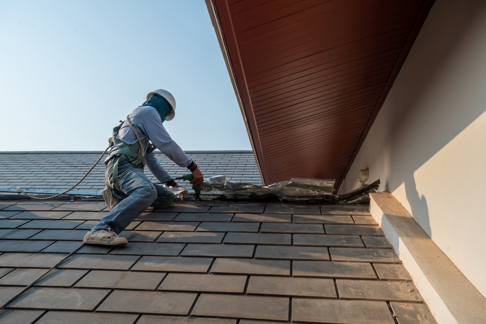 A construction worker wearing safety gear is repairing or installing shingles on a house roof. They are using tools to adjust the roofing material while being secured with a safety harness and rope. The roof has a modern, rectangular shingle design.