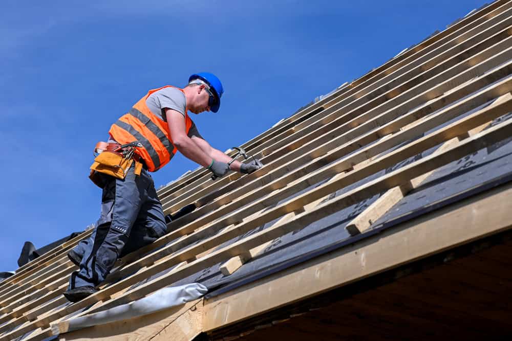 A construction worker wearing a blue hard hat and orange safety vest is nailing wooden beams on a roof, set against a clear blue sky.