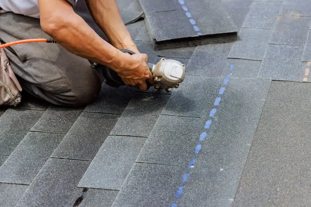 A person is using a nail gun to secure asphalt shingles on a roof. The person is wearing a gray shirt and gray pants, and the shingles are dark gray with blue guidelines.