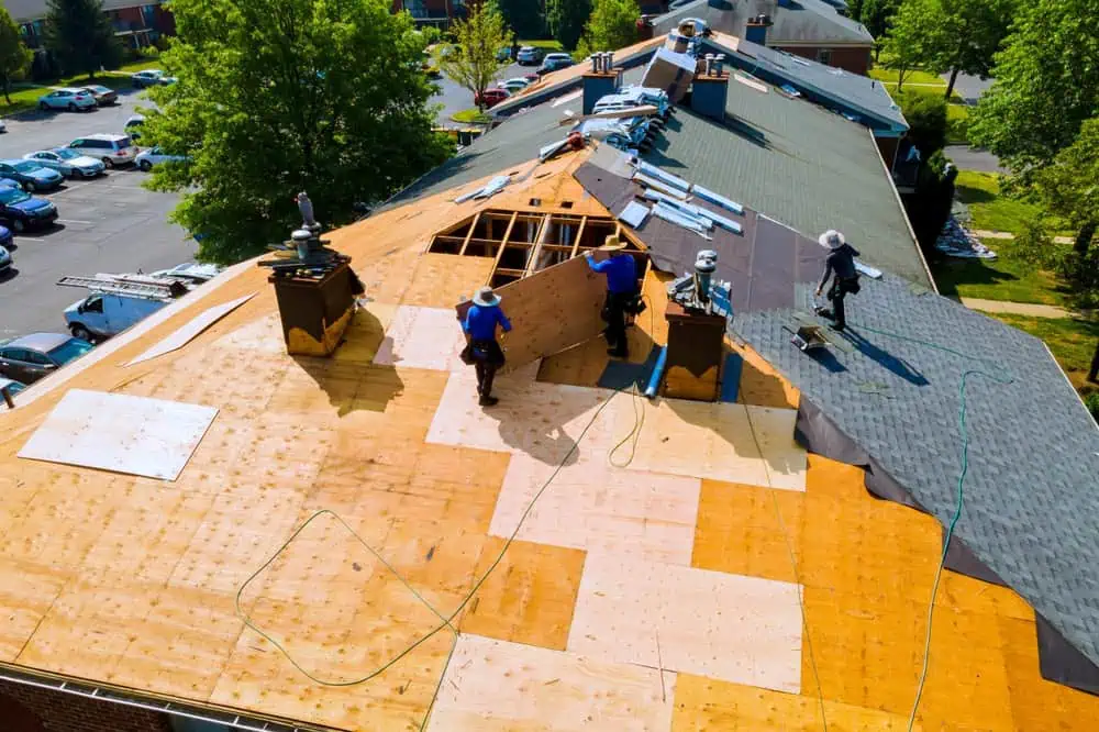 Aerial view of roofers working on a large building's roof. Sections of the roof are covered with wooden panels, and new shingles are being laid. Green trees and parked cars are visible in the background.