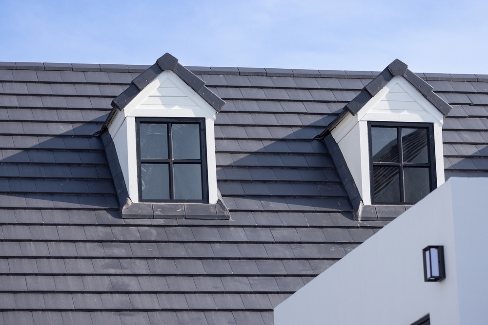 A house roof with dark gray shingles features two gabled dormer windows, each with four-pane black frames, against a clear blue sky. Part of a white exterior wall with a small rectangular light is visible in the foreground.