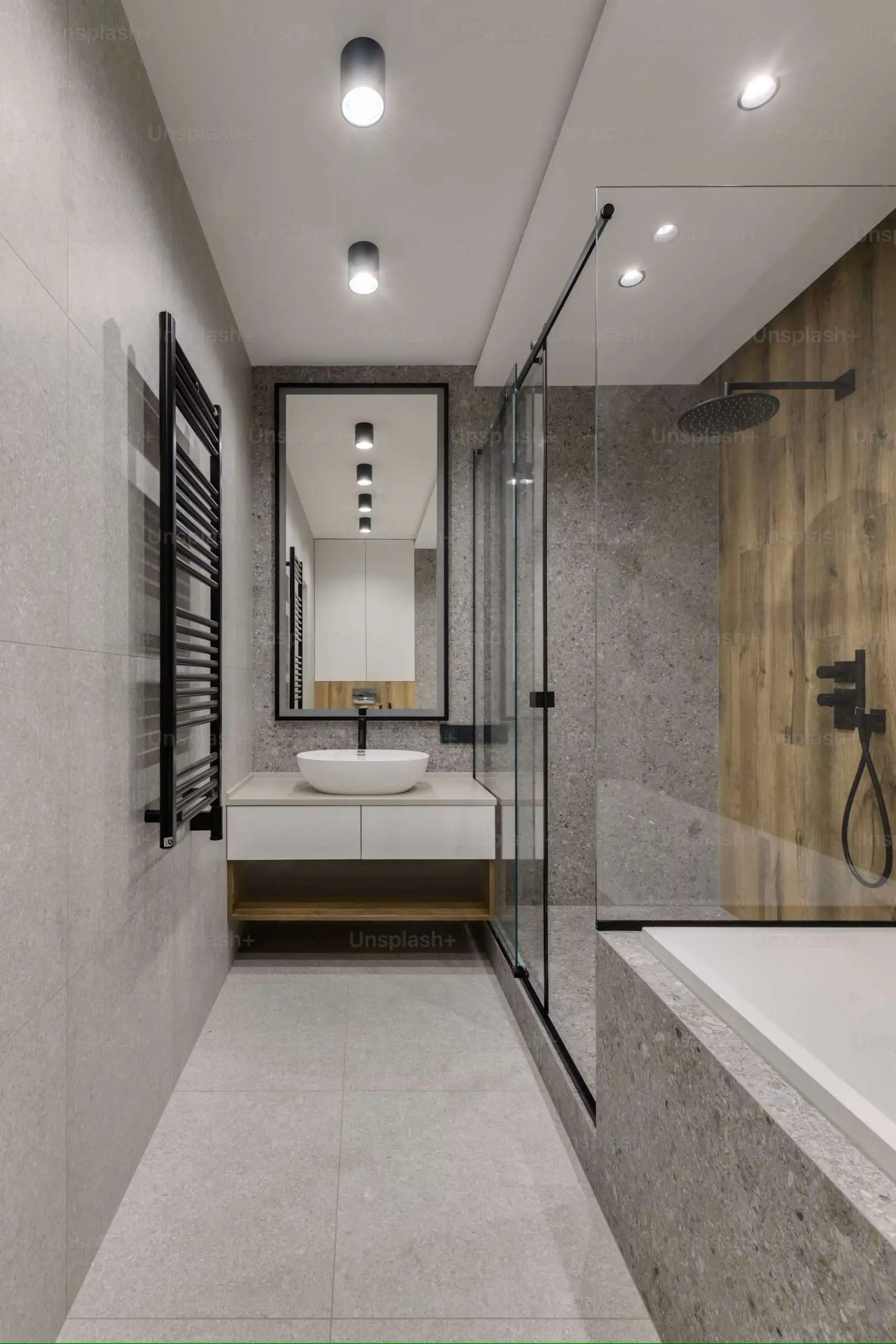 Modern bathroom with gray stone tiles, featuring a glass-enclosed shower with black fixtures, a wall-mounted sink with a round white basin, a tall mirror, and a heated towel rack on the left. Recessed ceiling lights provide illumination.