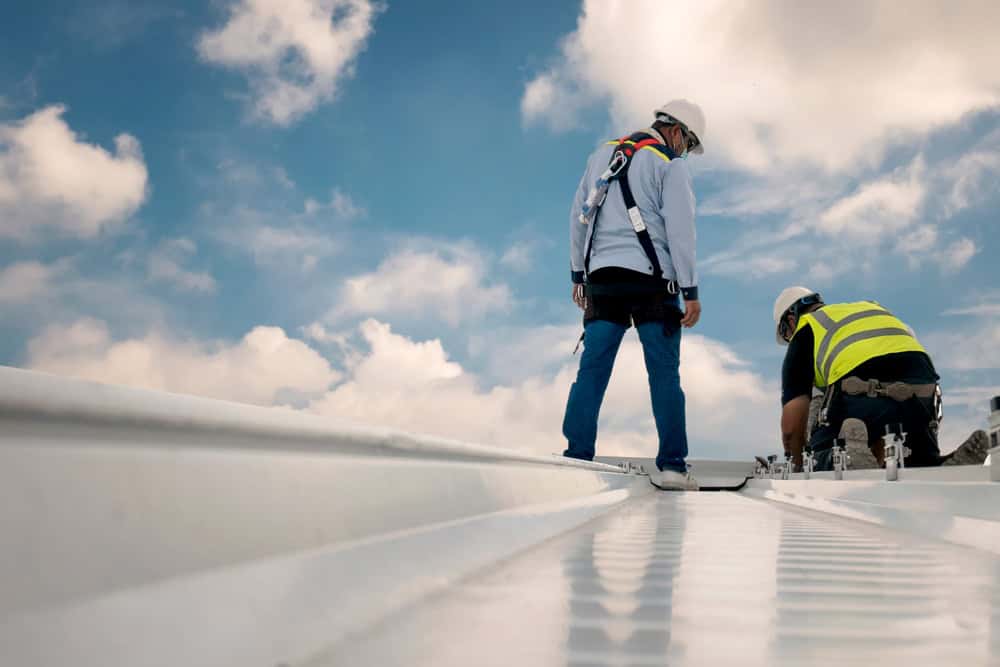 Two workers on a roof, wearing safety harnesses and helmets. One is kneeling, working on a task, while the other stands nearby. The sky is partly cloudy, creating a bright backdrop.