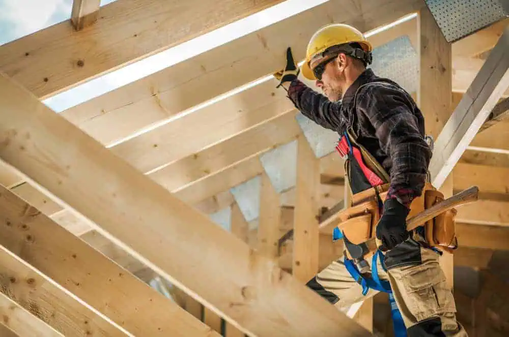 A construction worker wearing a yellow hard hat and safety harness works on a wooden roof frame. He holds a tool in one hand and braces a beam with the other, focused on building structure under a clear sky.