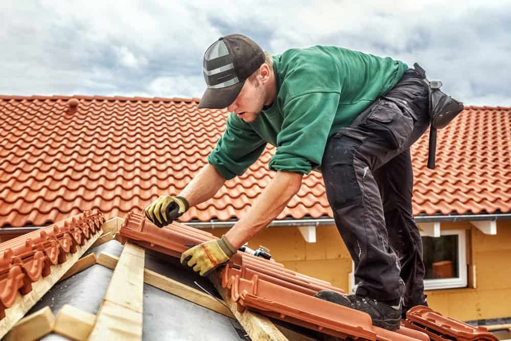 A construction worker in a green shirt and cap is laying terracotta roof tiles on a house. He is positioned on the roof ridge, wearing gloves and using tools, with a background of clear sky and completed tiled areas.