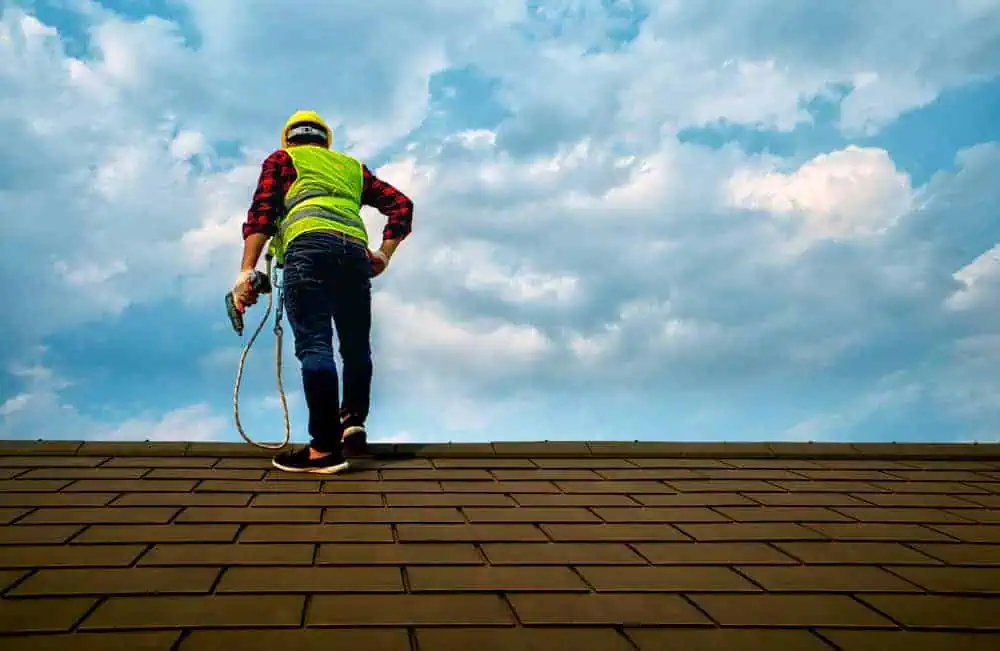 A construction worker in a yellow safety vest and helmet stands on a roof, holding a tool. The sky is partially cloudy in the background.