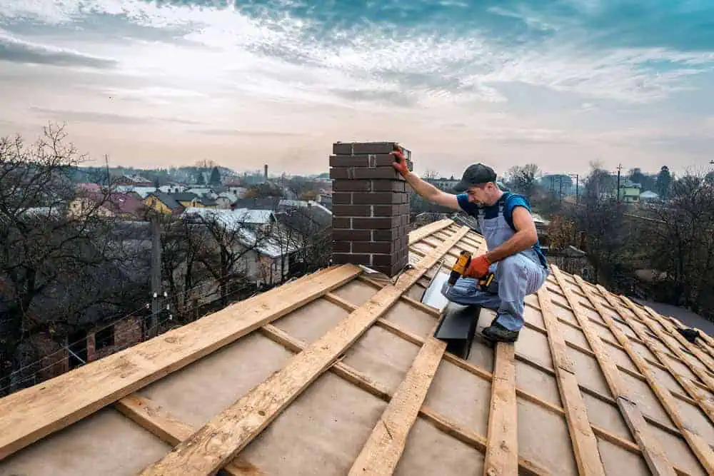 A construction worker installs bricks on a chimney atop a wooden roof frame. He is kneeling, using a tool, with a town visible in the background under a partly cloudy sky.