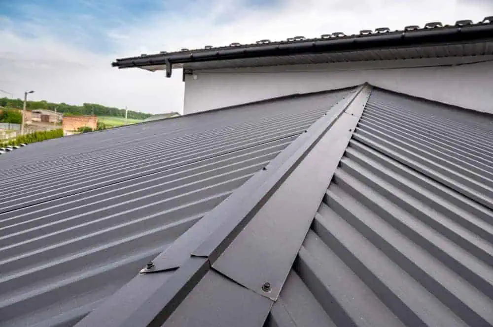 A close-up view of a modern, sloped metal roof with ribbed panels and ridge cap. The sky above is partly cloudy, and part of the building's gutter system is visible.