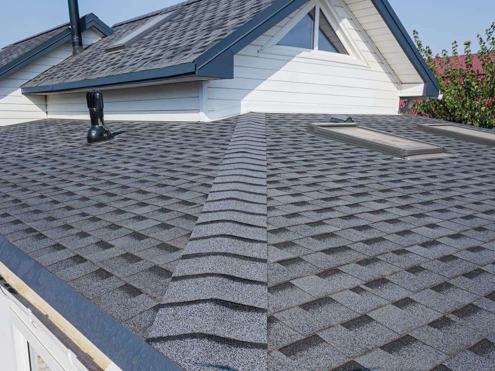 Close-up of a house roof with gray asphalt shingles. The roof features a skylight and a vent pipe. The house has white siding and a triangular window beneath the gable. A clear blue sky is visible in the background.