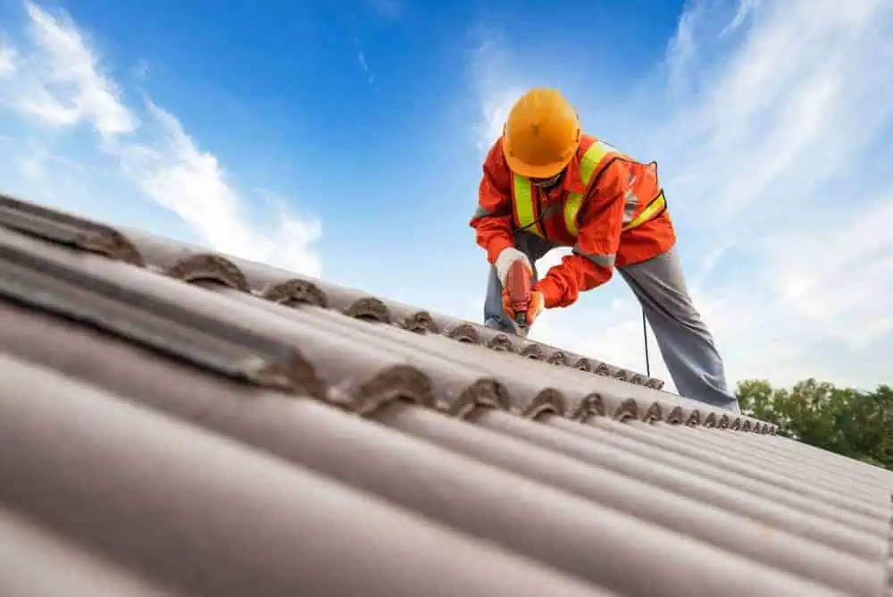 A construction worker in an orange safety vest and yellow hard hat is installing or repairing tiles on a rooftop. The sky is blue with a few clouds in the background. The worker is using a tool on the roof.