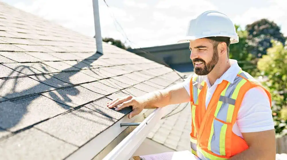 A construction worker wearing a white hard hat and orange safety vest inspects a roof. He is touching a shingle while holding a clipboard. The background shows trees and part of a building under a sunny sky.