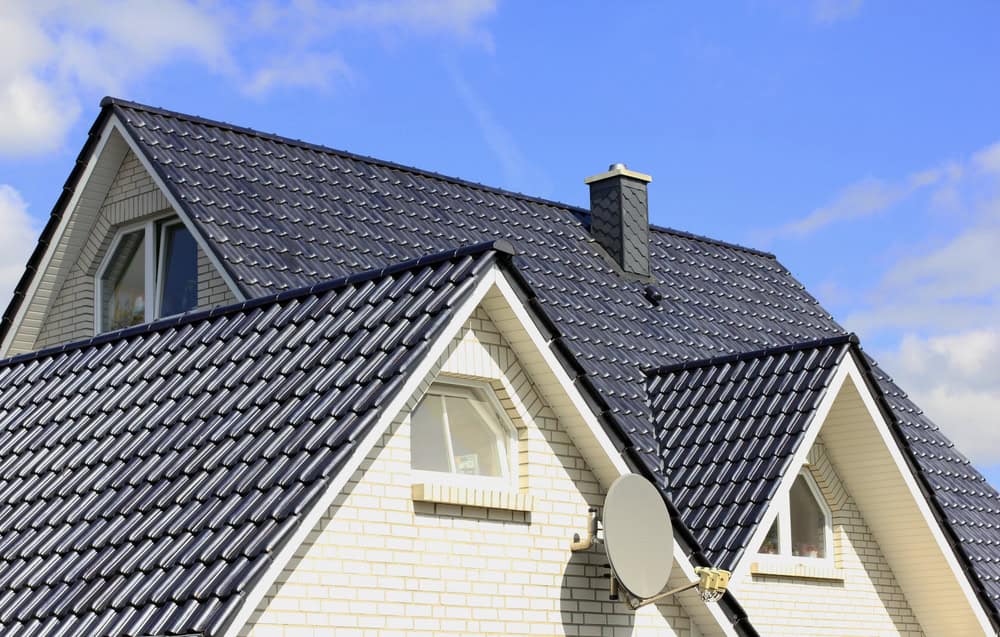 A white brick house with steep, dark gray tile roofing against a blue sky. The roof features multiple gables and a chimney. A satellite dish is attached to the wall below one of the arched windows.