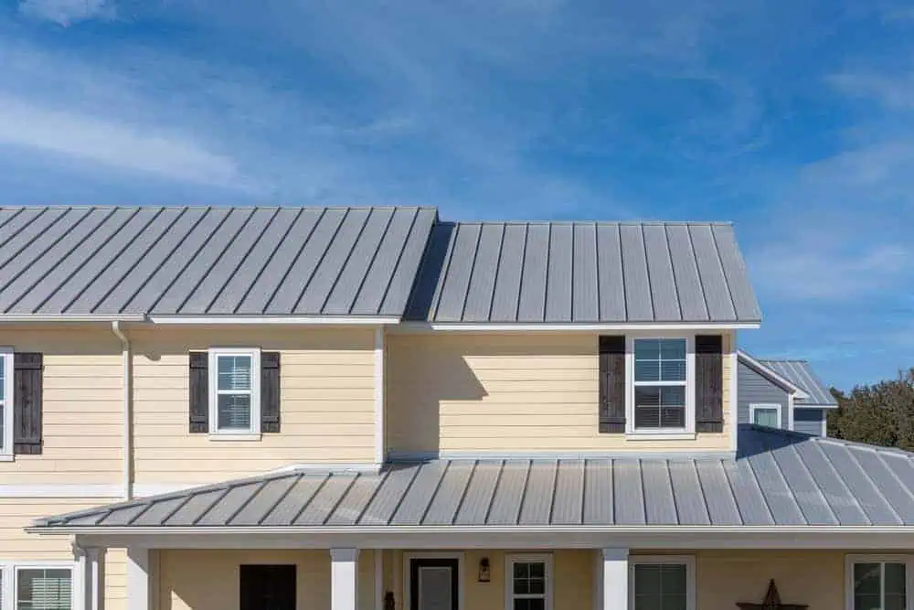 A two-story house with yellow siding and a gray metal roof under a clear blue sky. The facade features several windows with white frames and dark shutters.